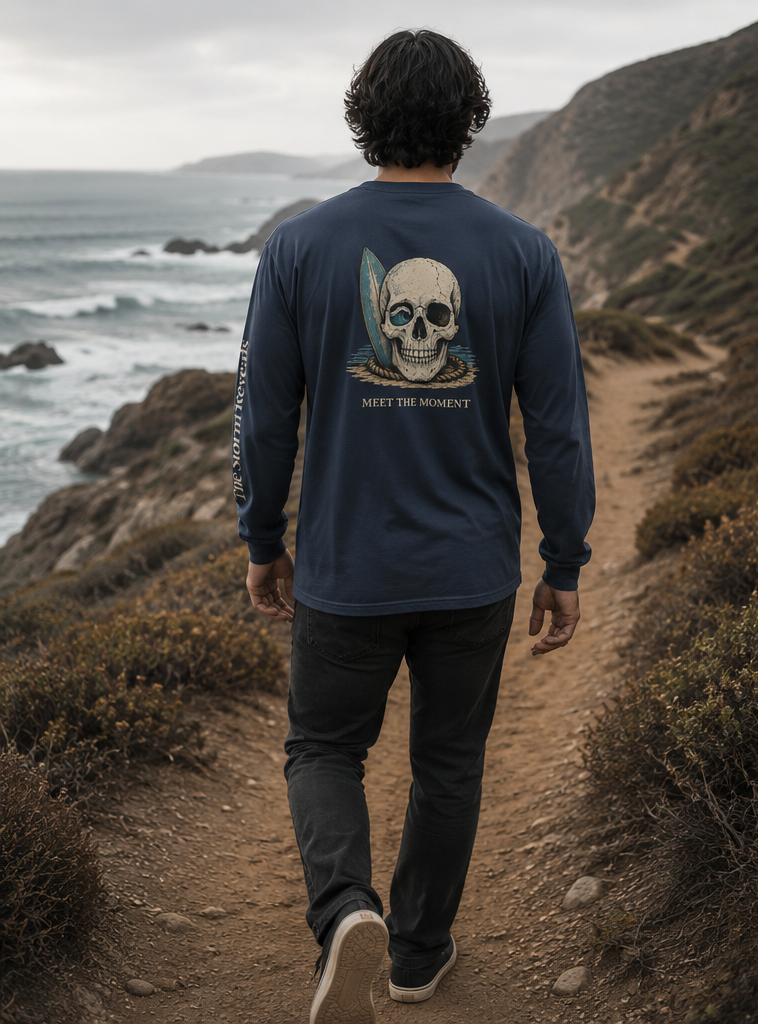 A man wearing a navy blue long-sleeve shirt with a large skull graphic and the text 'CATCH THE WAVE' on the back, shown in a split outdoor coastal scene. The left panel shows him from behind walking on a path, and the right panel shows him in side profile against the ocean.