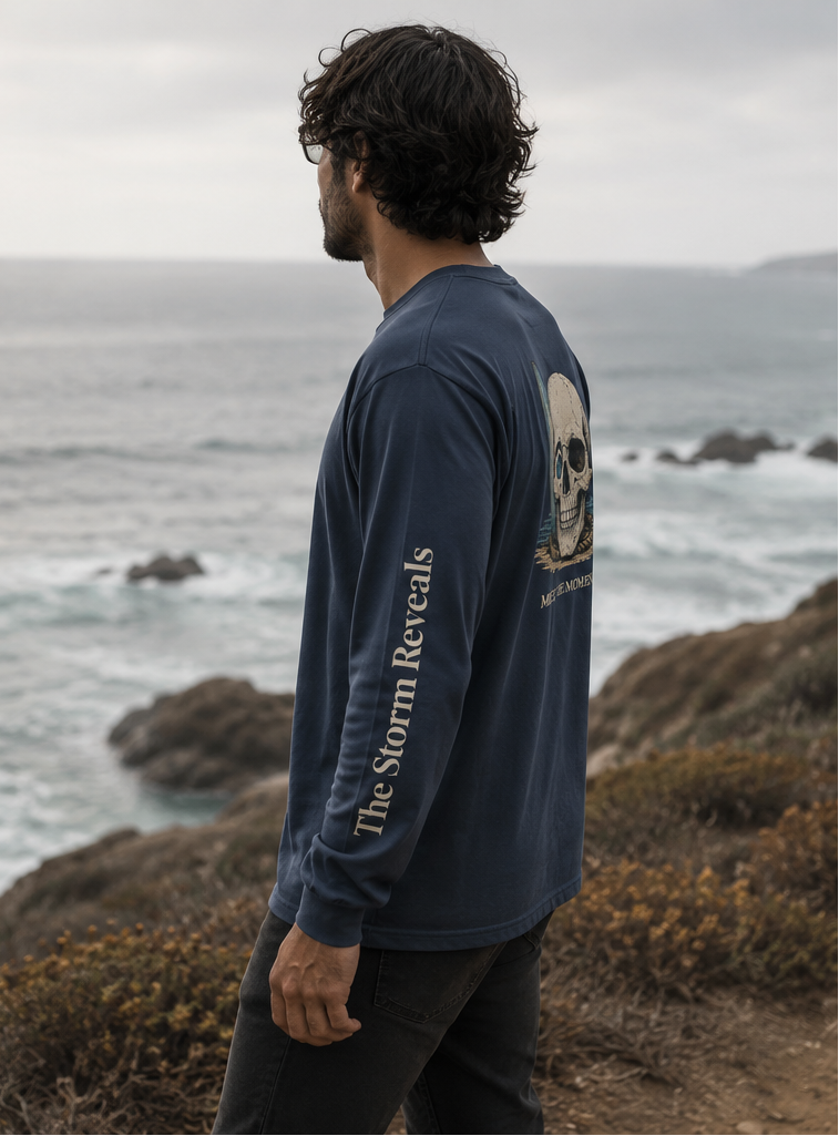 A man wearing a navy blue long-sleeve shirt with a large skull graphic and the text 'CATCH THE WAVE' on the back, shown in a split outdoor coastal scene. The left panel shows him from behind walking on a path, and the right panel shows him in side profile against the ocean.