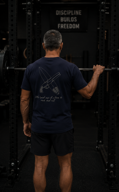 A man viewed from behind in a gym, wearing a navy blue t-shirt with a white graphic of a rifle and the text 'Discipline builds freedom' on the back, paired with black shorts.
