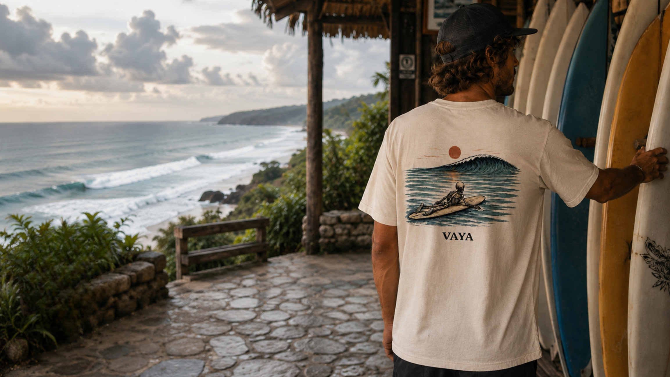A man wearing a cream-colored VAYA t-shirt with a wave graphic on the back, standing on a stone path next to surfboards with an ocean view in the background.